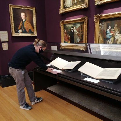 A highlight image for Person looking at a display of manuscripts. Paintings on the wall in the background.